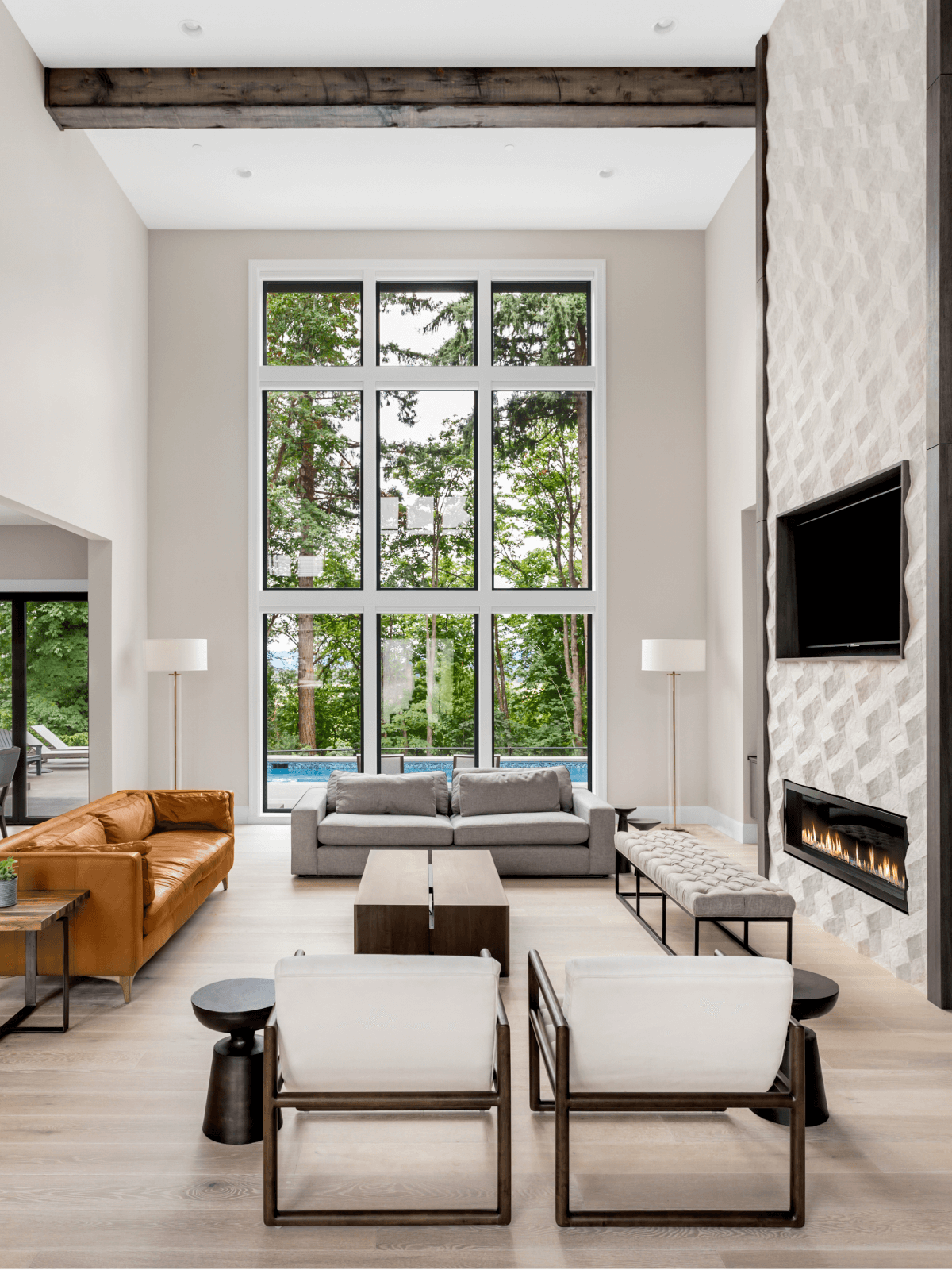 Beautiful living room in new luxury home wall of windows, floor to ceiling fireplace surround, hardwood floors, and exterior view of pool and trees.